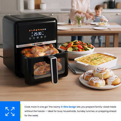 Black air fryer on a kitchen counter with various dishes, including vegetables and bread rolls.