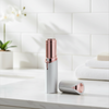 A handheld electric facial shaving device with a white body and rose gold metallic accents, shown standing upright with its cap placed beside it on a white marble countertop in a bright bathroom setting.