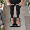 A person standing barefoot on a digital body scale in a bathroom, wearing black leggings. The scale has a black frame with gray platforms and a digital display showing 58.