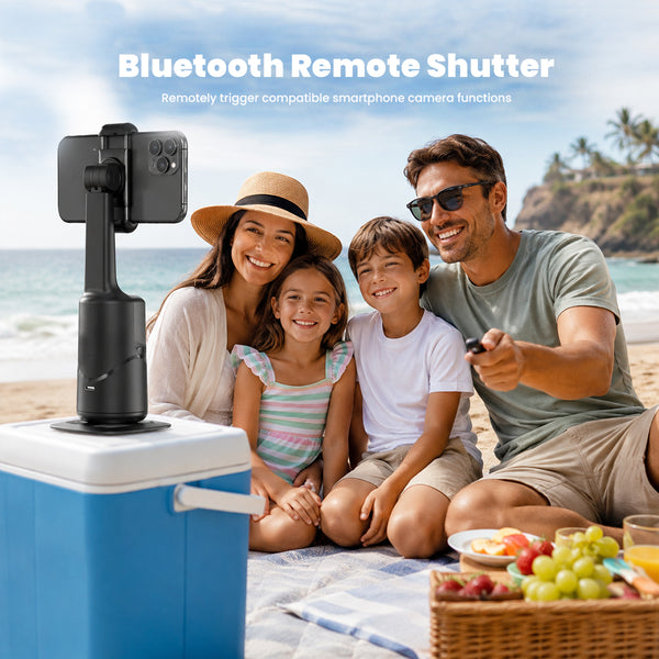 A family of four is sitting on a beach blanket on a sunny beach, using a Bluetooth remote shutter app to control a smartphone camera mounted on a black handheld gimbal. In the foreground, a blue and white cooler holds the camera setup.