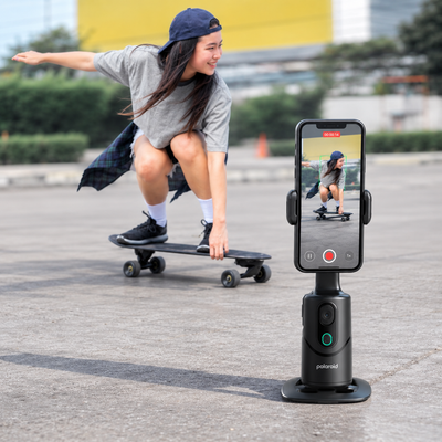 A woman skateboarding in the background while a Polaroid handheld phone holder in the foreground records her video call on a smartphone.